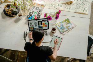 A small business owner works at her desk, with drawing pads and a laptop open.